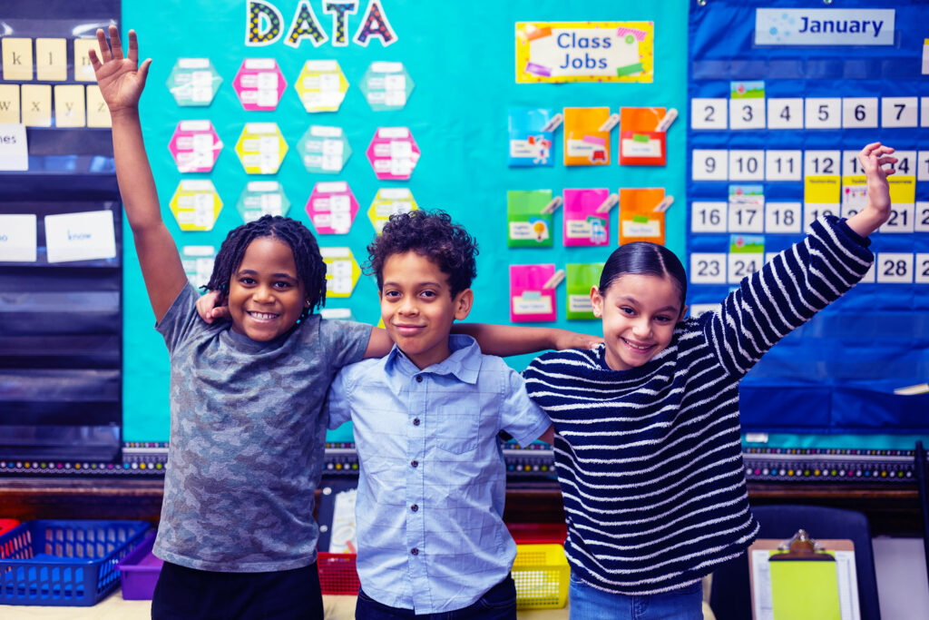 Smiling students with arm around each other and hands up in a classroom.